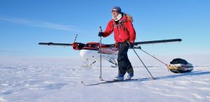 A person in winter gear skis across the snowy landscape of Antarctica, pulling a sled with supplies. An Antarctic Logistics and Expeditions red and white propeller plane on skis is parked in the background under a clear blue sky.
