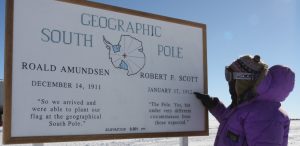 A person in a purple parka and goggles points at a large sign reading Geographic South Pole with expedition dates, quotes, and a map of Antarctica. Snow and blue sky set the scene for this Antarctic Logistics and Expeditions moment.