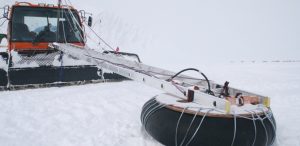 An orange snowcat vehicle in Antarctica, operated by Antarctic Logistics and Expeditions, pulls a long metal beam attached to a large black rubber tire, both connected with ropes and cables across the snowy landscape.