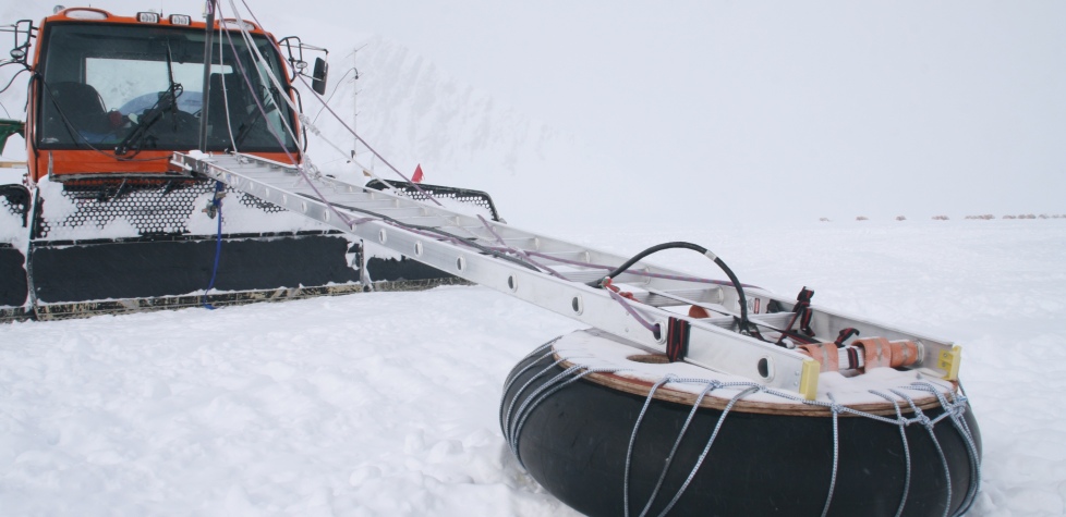 An orange snowcat vehicle in Antarctica, operated by Antarctic Logistics and Expeditions, pulls a long metal beam attached to a large black rubber tire, both connected with ropes and cables across the snowy landscape.