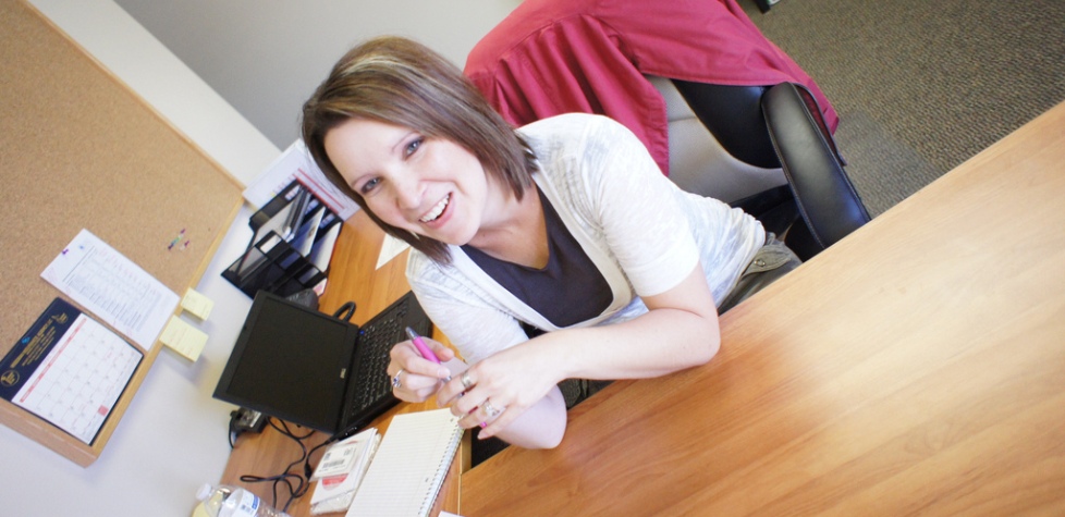 A woman with short brown hair sits at a wooden desk in an office, smiling up at the camera. A calendar, notebook, pens, and laptop are on the desk—a typical day at Antarctic Logistics and Expeditions as she plans upcoming work in Antarctica.