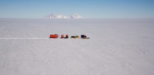 A convoy of red and yellow vehicles from Antarctic Logistics and Expeditions travels across a vast, snowy expanse in Antarctica, with distant snow-covered mountains under a clear blue sky.
