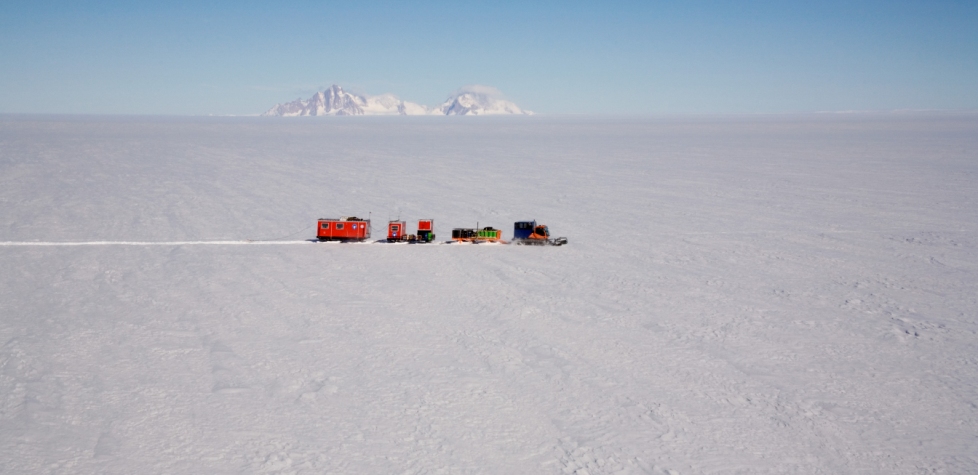 A convoy of red and yellow vehicles from Antarctic Logistics and Expeditions travels across a vast, snowy expanse in Antarctica, with distant snow-covered mountains under a clear blue sky.