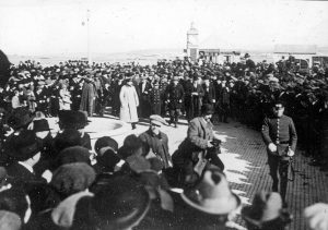 A large crowd of people, many wearing hats and coats, gathers outdoors on a paved area; some uniformed individuals are present, and a domed building reminiscent of the South Pole stands in the background.
