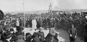 A large crowd, many in hats and coats, gathers outdoors around a circular area—evoking the spirit of historic Antarctic Logistics and Expeditions. In the background stand a clock tower and open land; a uniformed officer stands in the foreground.