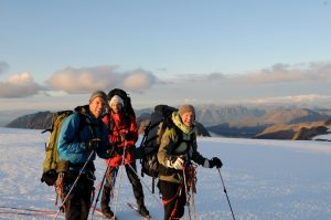 Three climbers in winter gear and backpacks smile on a snowy mountain slope with ski poles. The sun is low over distant mountains and clouds under a clear sky—an epic moment during their Antarctic Logistics and Expeditions adventure.