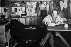 Black-and-white photo of a man sitting at a desk in a wooden cabin, writing in a book. In this Antarctic setting, shelves with books, papers, and hanging clothes are visible. A bed, mug, and scattered items add to the cozy South Pole room.