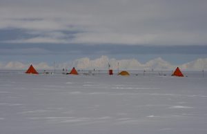 A row of small orange tents and equipment are set up on a vast, snow-covered plain in Antarctica under a cloudy sky, with distant mountains visible on the horizon—an expedition scene typical of Antarctic Logistics and Expeditions near the South Pole.