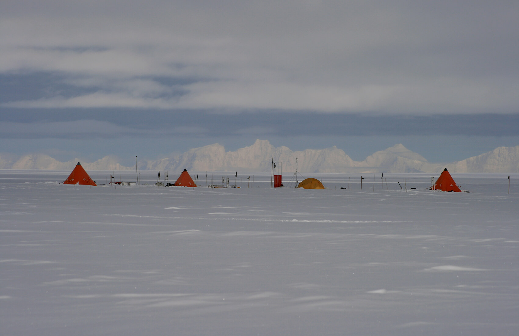 A row of small orange tents and equipment are set up on a vast, snow-covered plain in Antarctica under a cloudy sky, with distant mountains visible on the horizon—an expedition scene typical of Antarctic Logistics and Expeditions near the South Pole.