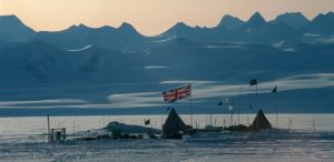 A snowy encampment with several tents and flags, including a large Union Jack, stands in flat, icy Antarctica. Supported by Antarctic Logistics and Expeditions, it lies beneath a pale sky with distant, jagged mountains in view.