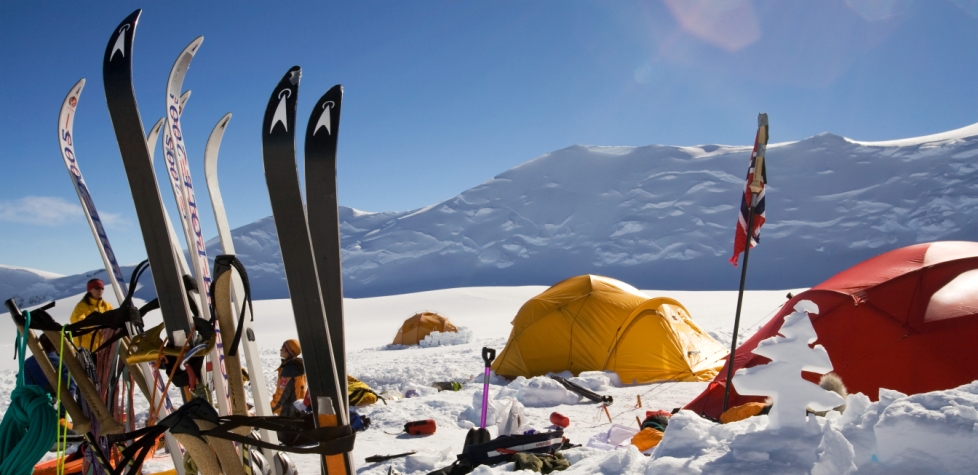Ski equipment stands in the snow near bright tents at an Antarctic Logistics and Expeditions campsite on a snowy mountain in Antarctica, with people in winter gear and snow-covered peaks beneath a clear blue sky.