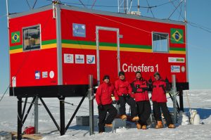 Four people in red jackets stand in front of a red research module labeled Criosfera 1 with Brazilian flags on it, on a snowy Antarctic landscape under a clear blue sky, highlighting scientific efforts supported by Antarctic Logistics and Expeditions.