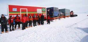 A group of people in heavy winter clothing stand beside a large red and green mobile research station with a Brazilian flag, set on the snowy landscape of Antarctica, with Antarctic Logistics and Expeditions vehicles and trailers in the background.