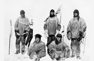 Five explorers in heavy cold-weather gear pose on snowy ground at the South Pole with flags behind them, against a blank, icy backdrop. Two sit in front, three stand behind, all looking toward the camera.