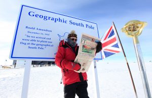 A person in a red winter coat stands at the Geographic South Pole in Antarctica, holding a newspaper. Behind them is a large sign, a British flag, and the ceremonial South Pole marker, all set in a snowy landscape.