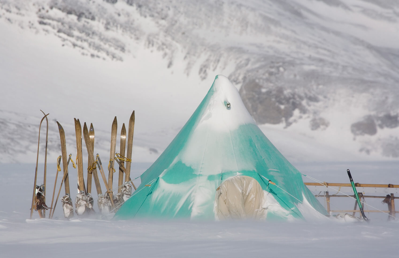 A green tent covered in snow stands in a snowy Antarctic landscape, surrounded by skis and poles upright in the snow, with the majestic mountains of Antarctica in the background.
