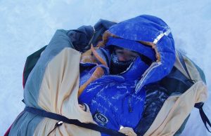 A person bundled in heavy winter clothing and a blue hooded jacket lies wrapped in blankets on snow near the South Pole, with frost visible on their face and gear.