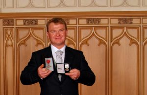A man in a suit smiles and holds up two open medal boxes with medals inside, standing before a wooden paneled wall with decorative arches—recognizing achievements related to Antarctic Logistics and Expeditions.