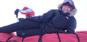 A person in winter gear smiles while lying on a red sled in the snowy environment of Antarctica. Another, outfitted by Antarctic Logistics and Expeditions, is seen in the background adjusting equipment.