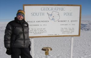 A person in winter clothing stands next to a large South Pole sign in Antarctica, with snow and ice stretching behind. The sign honors Roald Amundsen and Robert F. Scott’s historic Antarctic expeditions.