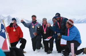 Six people wearing winter gear and medals kneel and stand on snow in Antarctica, smiling and holding glasses. Snowy mountains are in the background, a flag is visible on the left, celebrating an Antarctic Logistics and Expeditions achievement.