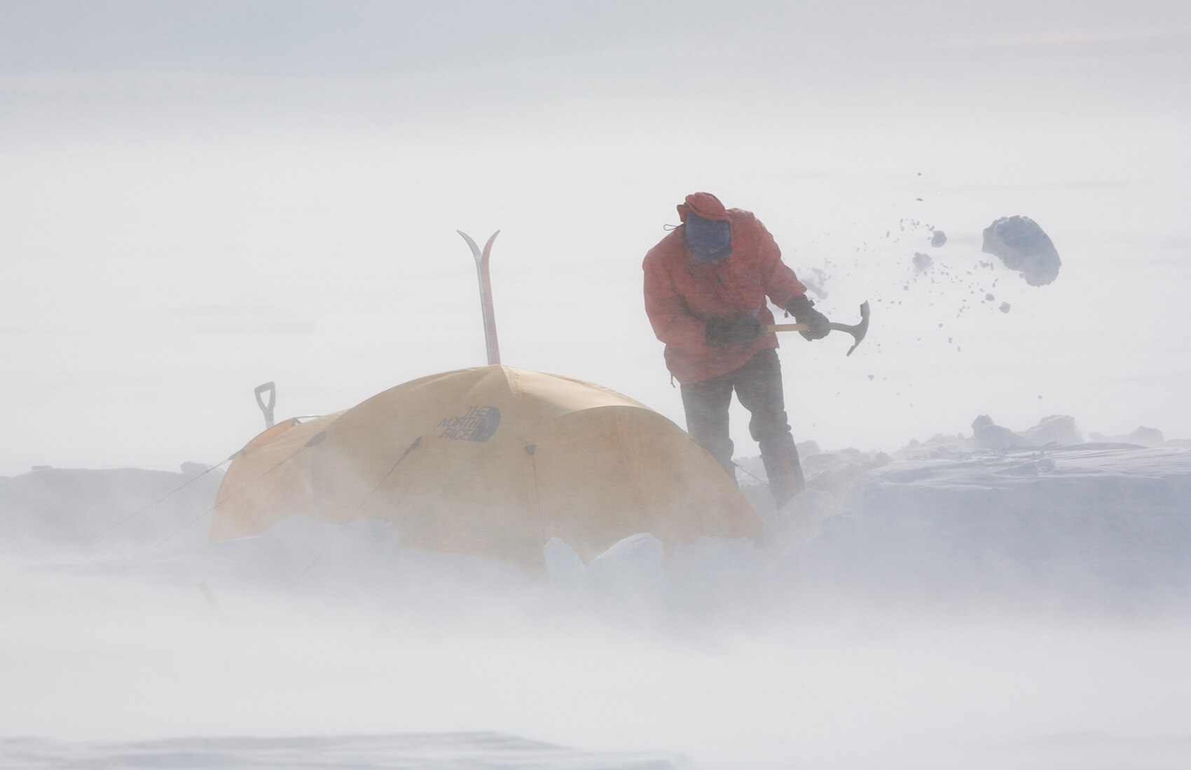 A person in winter clothing uses an ice axe to clear snow around a yellow tent in Antarctica, with snow swirling in the air—capturing the spirit of Antarctic Logistics and Expeditions near the South Pole.