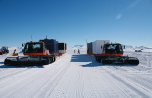 Two orange snow vehicles with large treads transport container units across a snowy landscape in Antarctica under a clear blue sky, with mountains and a person visible in the background—an example of Antarctic Logistics and Expeditions at work.