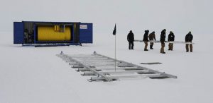 A group of people in winter clothing carry a long piece of equipment across a snowy Antarctic landscape, near a blue shipping container and metal structures, as part of Antarctic Logistics and Expeditions operations.