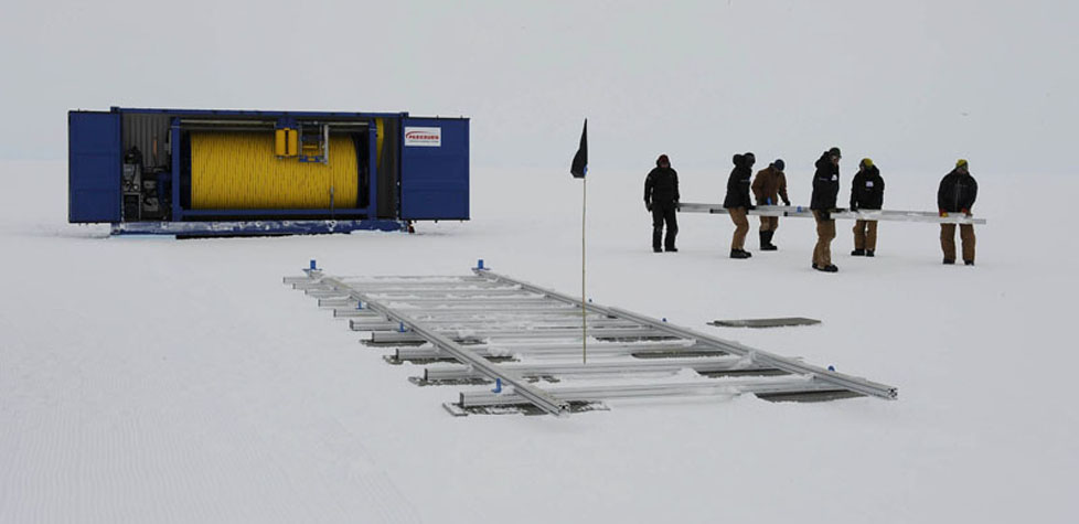 A group of people in winter clothing carry a long piece of equipment across a snowy Antarctic landscape, near a blue shipping container and metal structures, as part of Antarctic Logistics and Expeditions operations.