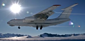 A large white cargo plane flies low over a snowy, mountainous Antarctic landscape with the sun shining brightly behind it—a stunning scene of Antarctic Logistics and Expeditions under a clear blue sky with some lens flare.