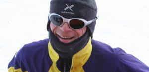 A person dressed in winter gear, including a grey hat, white sunglasses, and a yellow and purple jacket, smiles while standing outdoors on snow during an Antarctic Logistics and Expeditions adventure.