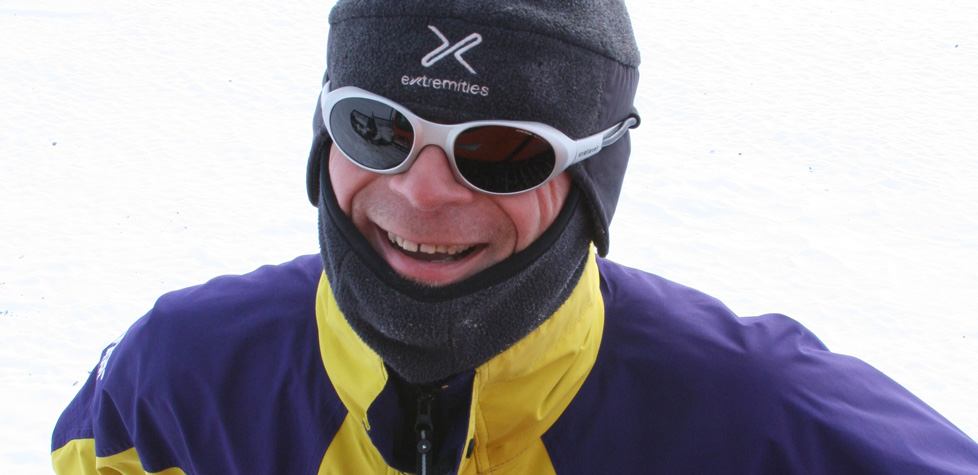 A person dressed in winter gear, including a grey hat, white sunglasses, and a yellow and purple jacket, smiles while standing outdoors on snow during an Antarctic Logistics and Expeditions adventure.