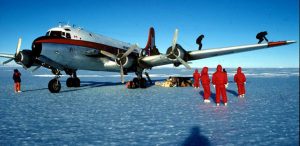 A group of people in red cold-weather suits work on a snowy, icy surface in Antarctica next to a large propeller airplane, with some individuals on top of the aircraft under a clear blue sky.