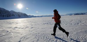 A person in a red jacket runs across a snow-covered landscape with mountains in the background, under a bright sun and clear blue sky—capturing the spirit of Antarctic adventure supported by Antarctic Logistics and Expeditions.