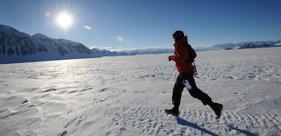 A person in a red jacket runs across a snow-covered landscape with mountains in the background, under a bright sun and clear blue sky—capturing the spirit of Antarctic adventure supported by Antarctic Logistics and Expeditions.