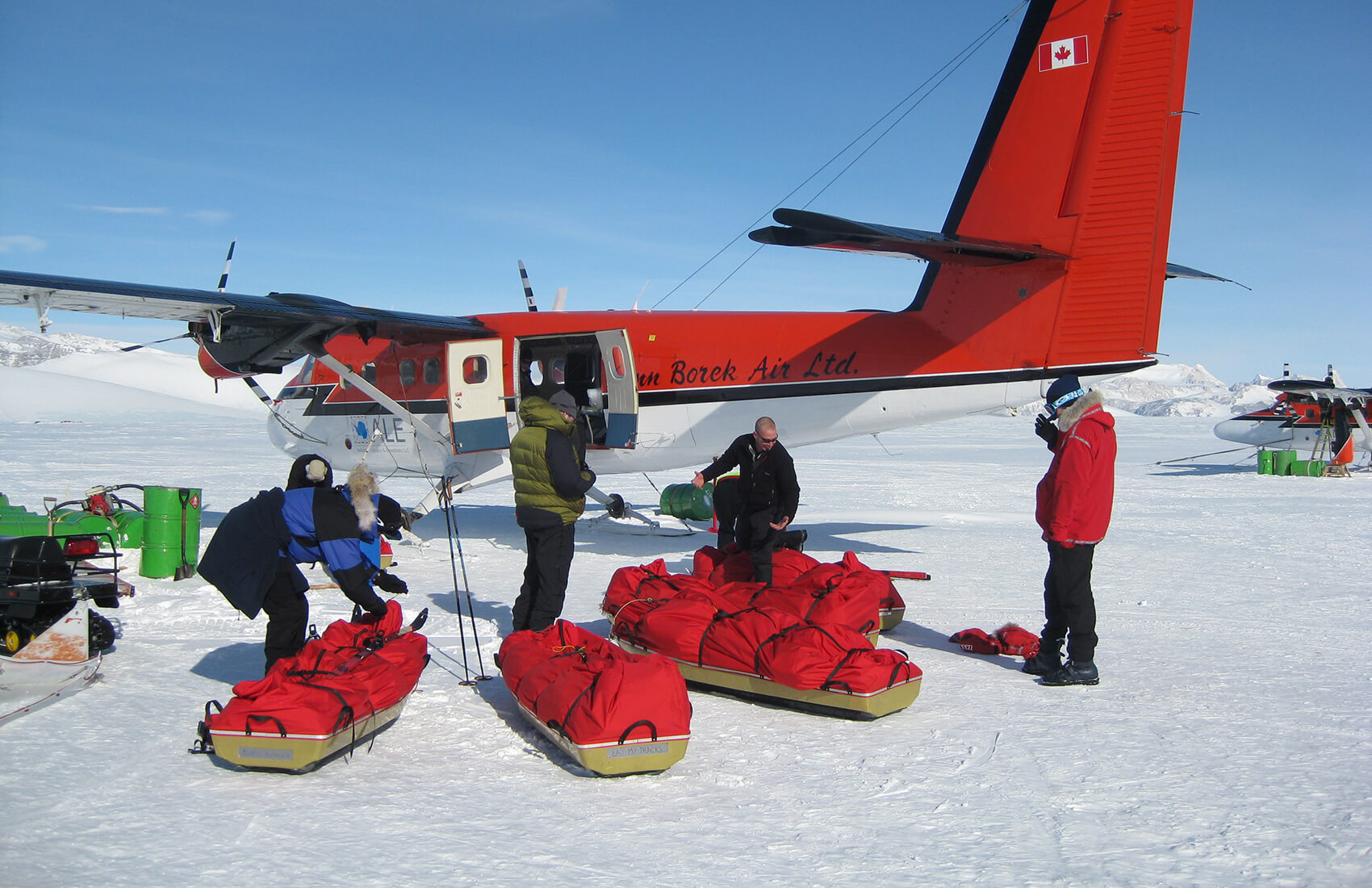Four people in winter gear load red supplies onto sleds beside an orange and white Twin Otter airplane in Antarctica, with another plane and Antarctic Logistics and Expeditions’ supplies visible on the snowy terrain in the background.
