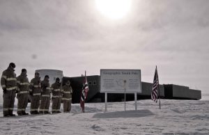 Seven people in heavy cold-weather gear stand by a sign reading Geographic South Pole, with American and Norwegian flags on either side, and a large building in snowy Antarctica under a bright sun—an adventure made possible by Antarctic Logistics and Expeditions.