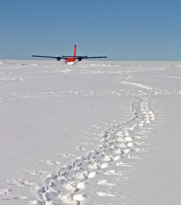 A red and white small airplane, operated by Antarctic Logistics and Expeditions, is parked on a vast, snow-covered landscape in Antarctica under a clear blue sky, with winding footprints leading from the foreground toward the plane.