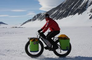 A person wearing a red jacket, black pants, and sunglasses rides a fat-tire bike on snow with green panniers and gear in Antarctica. Snowy mountains and a blue sky with clouds create a stunning South Pole backdrop.