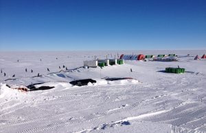 A snowy research outpost in Antarctica, supported by Antarctic Logistics and Expeditions, features green crates, tents, and equipment scattered across a flat, icy landscape under a clear blue South Pole sky.