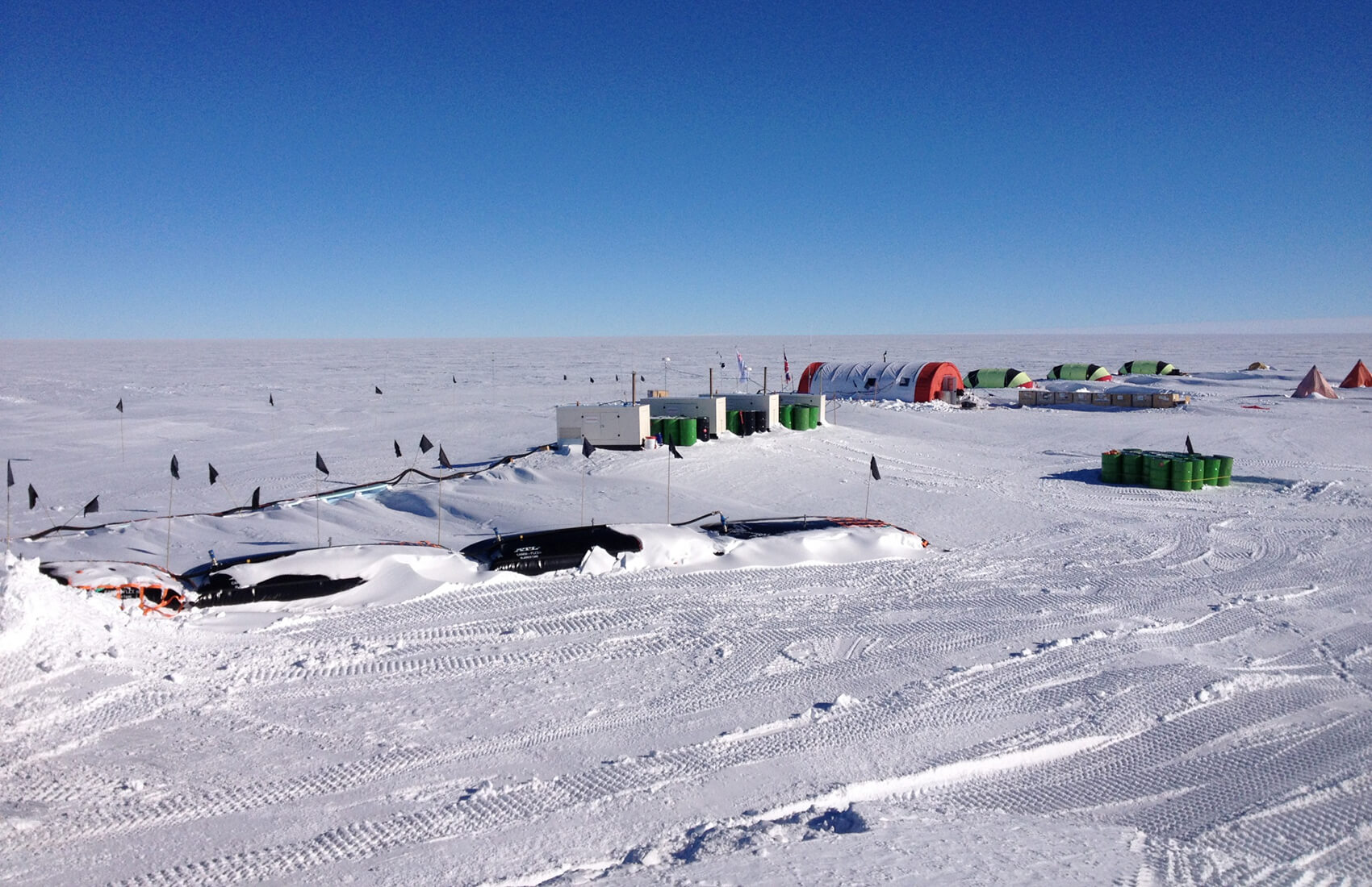 A snowy research outpost in Antarctica, supported by Antarctic Logistics and Expeditions, features green crates, tents, and equipment scattered across a flat, icy landscape under a clear blue South Pole sky.
