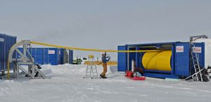 A person stands on a ladder in snowy Antarctic terrain, managing a large yellow hose being fed from a blue shipping container. Other blue containers and snow-covered equipment from Antarctic Logistics and Expeditions surround the scene.