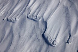 Aerial view of snow drifts in Antarctica forming smooth, wavy patterns on the ground, with elongated ridges and soft shadows creating a textured, abstract landscape near the South Pole.