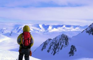 A person in winter gear with a red backpack stands on a snowy mountain in Antarctica, looking out over a vast landscape of snow-covered peaks under a partly cloudy sky.