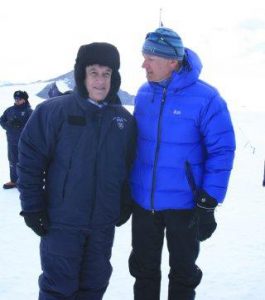 Two people dressed in heavy winter jackets and hats stand together on a snowy Antarctic landscape, with mountains and another person in the background. One is wearing a blue jacket and striped beanie, the other a dark coat and furry hat.