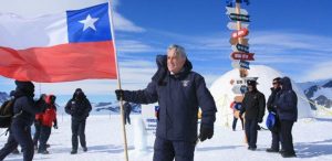 A man holds the Chilean flag on a snowy landscape in Antarctica, surrounded by several people. In the background, a dome structure from Antarctic Logistics and Expeditions and a colorful signpost stand under the clear blue sky.