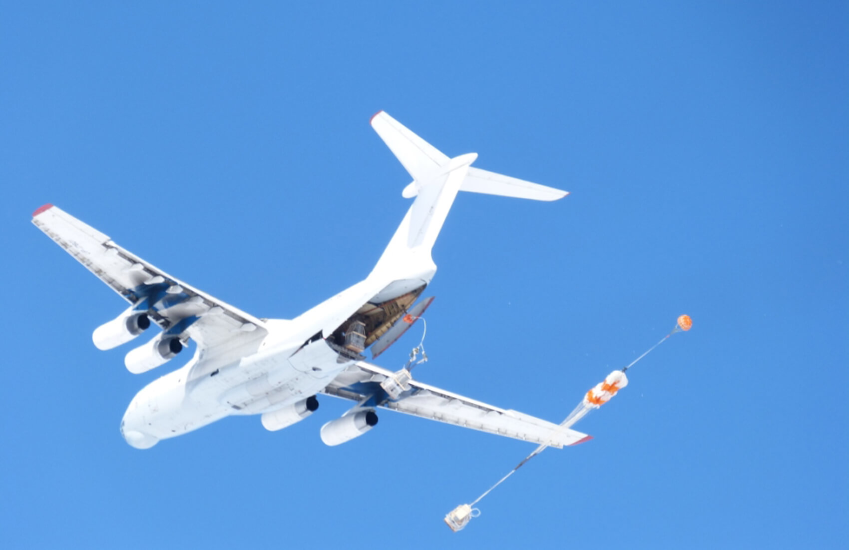A white cargo plane flies against a clear blue sky over Antarctica with its rear door open, deploying a parachute that is attached to a load being airdropped for Antarctic Logistics and Expeditions.