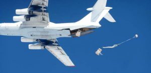 A large cargo airplane with Antarctic Logistics and Expeditions in flight drops several supply crates attached to parachutes from its open cargo bay against the clear blue sky over Antarctica.