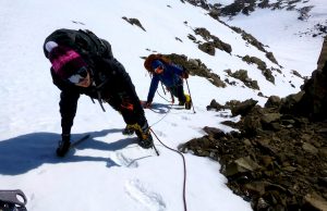 Two climbers in winter gear, helmets, and sunglasses ascend a steep, snowy Antarctic slope using ice axes and crampons, connected by a safety rope, with rocky terrain nearby—an adventure inspired by Antarctic Logistics and Expeditions.