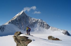 Two skiers stand on a snowy rocky ledge overlooking a vast, snow-covered mountain landscape in Antarctica, with a sharp, prominent peak under a clear blue sky—an adventure made possible by Antarctic Logistics and Expeditions.